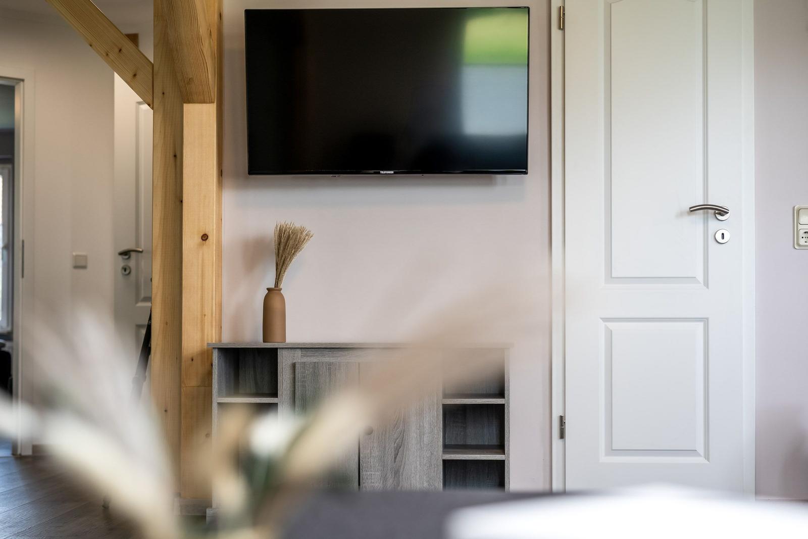Wandmontierte TV-Anlage über einem Holzregal mit Vase. Weiße Tür und Holzstütze sichtbar.