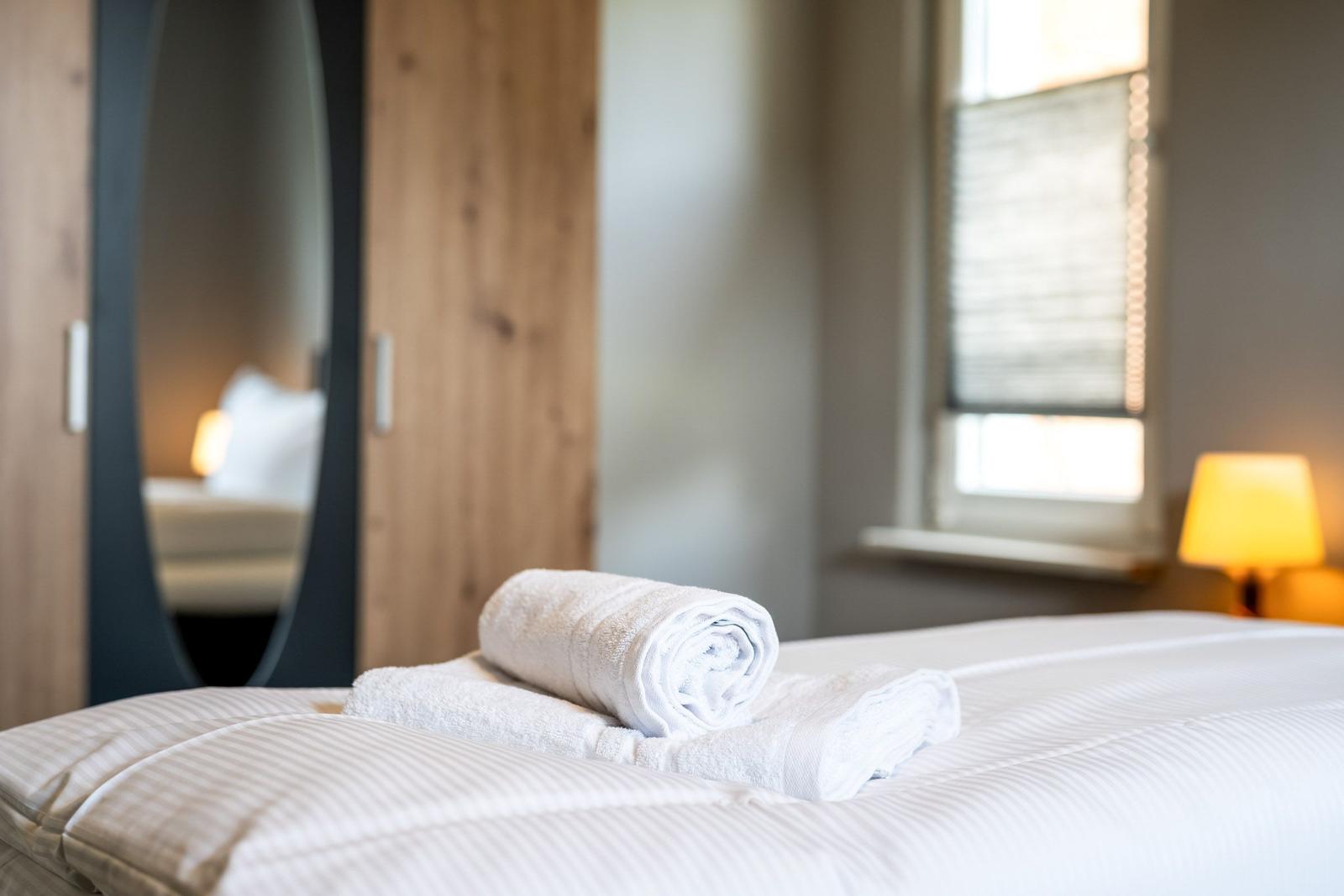 Ein Bett mit weißen Laken und gefalteten Handtüchern. Im Hintergrund ein Spiegel und ein Fenster.