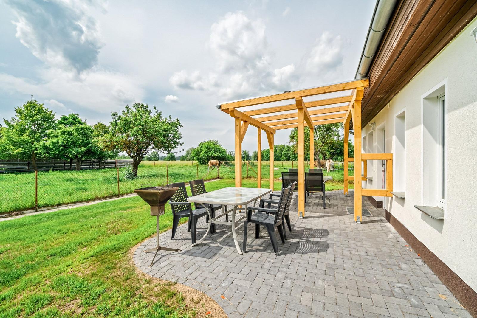 Terrasse mit Holzüberdachung, Tisch, Stühlen und Grill. Grünflächen und Zaun im Hintergrund.