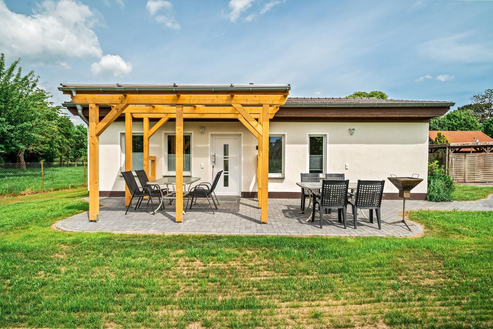 Terrasse mit Holzüberdachung, Tisch, Stühle und Grill. Haus mit Fenstern und Tür.