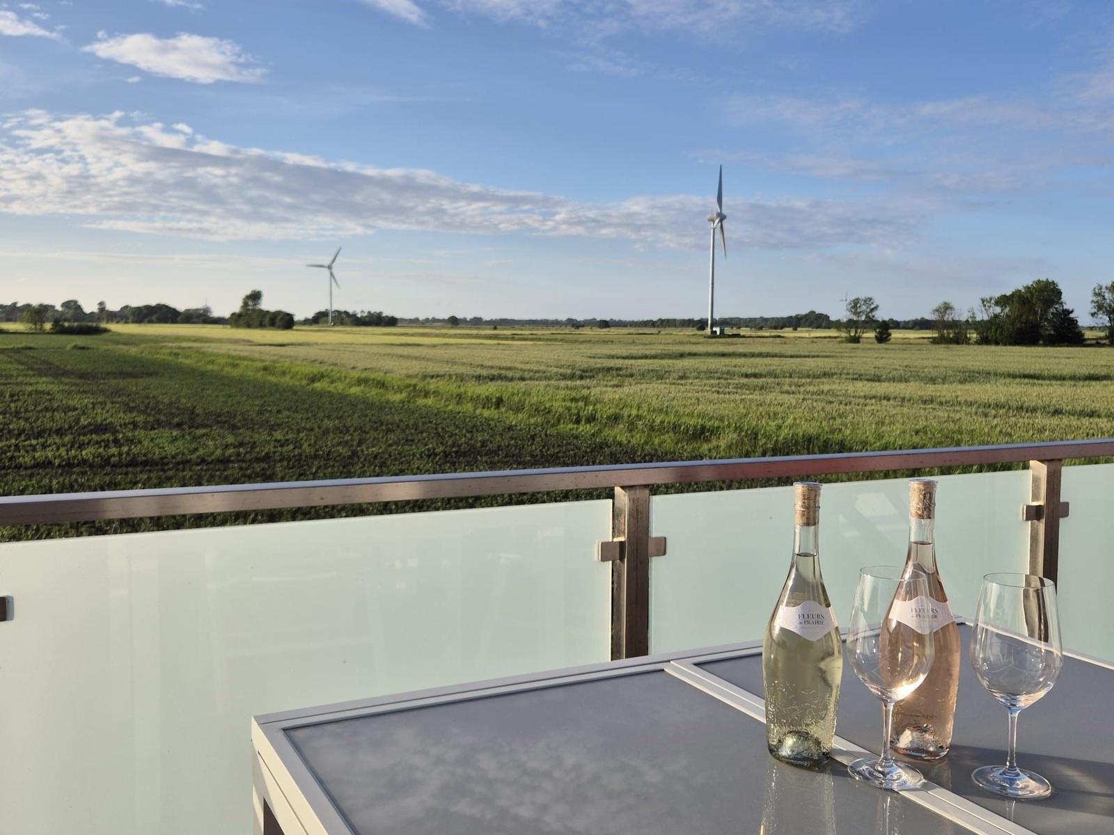Terrasse mit Tisch, zwei Weingläsern und Flaschen. Blick auf Felder und Windräder unter blauem Himmel.