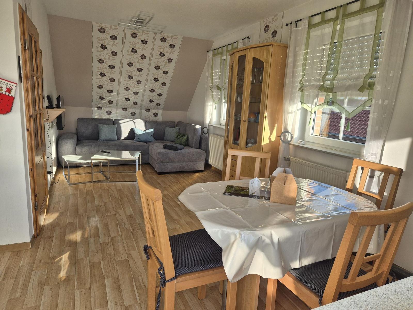 Wohn- und Essbereich mit Sofa, Esstisch und Holzmöbeln. Holzfußboden und Fenster mit Gardinen.