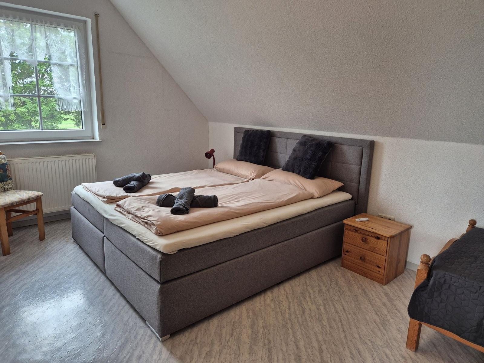 Doppelbett mit zwei Kissen, Nachttisch und Fenster mit Blick auf Grün.