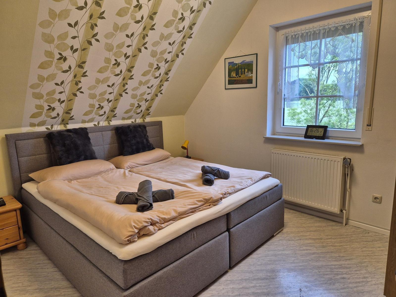 Schlafzimmer mit Doppelbett, Fenster und Wandtapete mit Blattmuster.