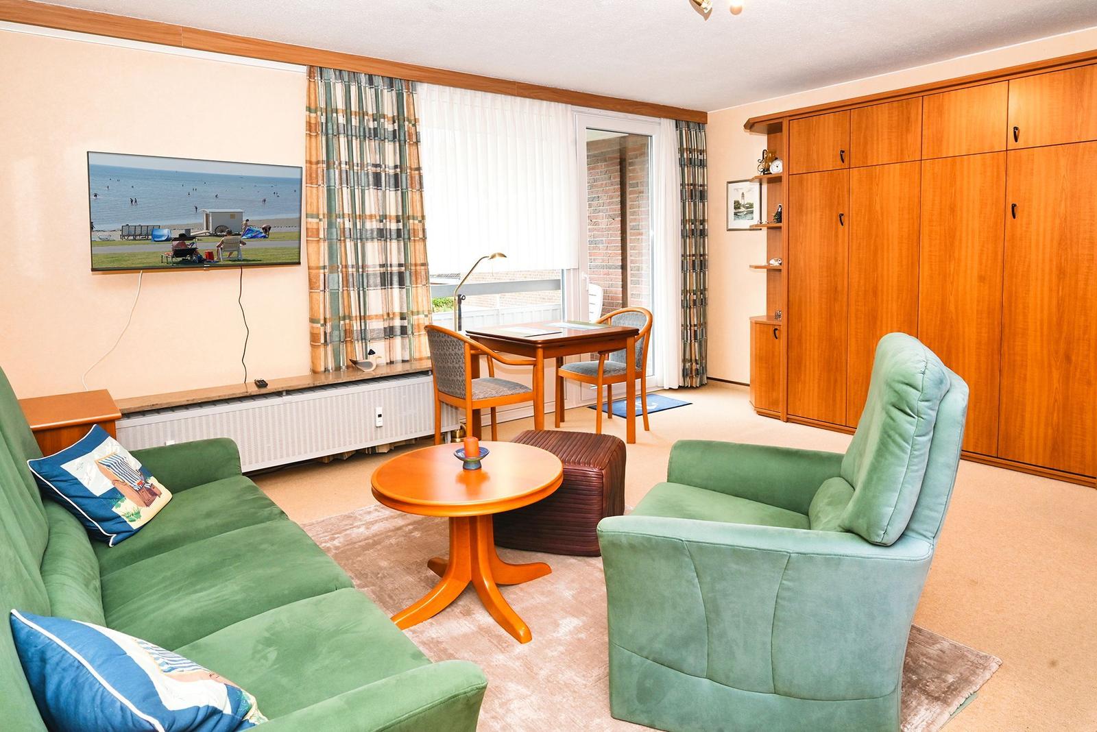 Wohnzimmer mit grüner Couch, Sessel, Tisch und Fernseher. Große Fenster mit Blick auf Balkon.