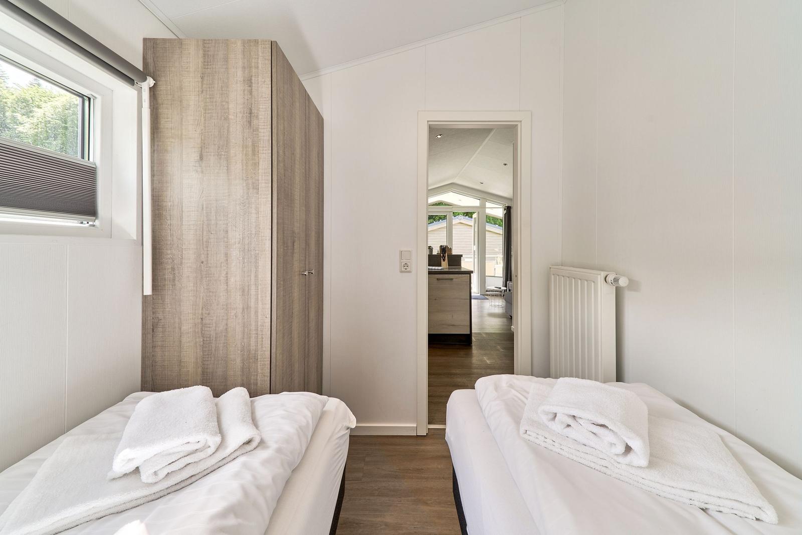 Zwei Betten mit weißen Laken und Handtüchern. Holzschrank und Fenster. Blick in die Küche.
