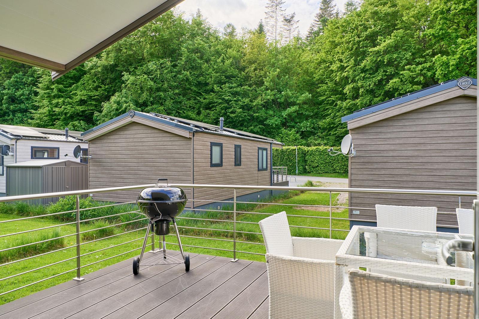 Terrasse mit Grill und Sitzgelegenheiten, Blick auf Ferienhäuser und grüne Bäume.