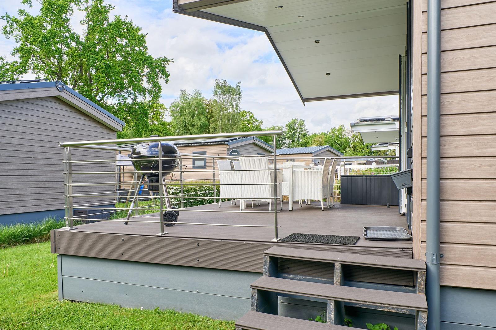 Terrasse mit Grill, weißem Gartenmöbel und Holzstufen. Hintergrund: Wohnwagen und Bäume.