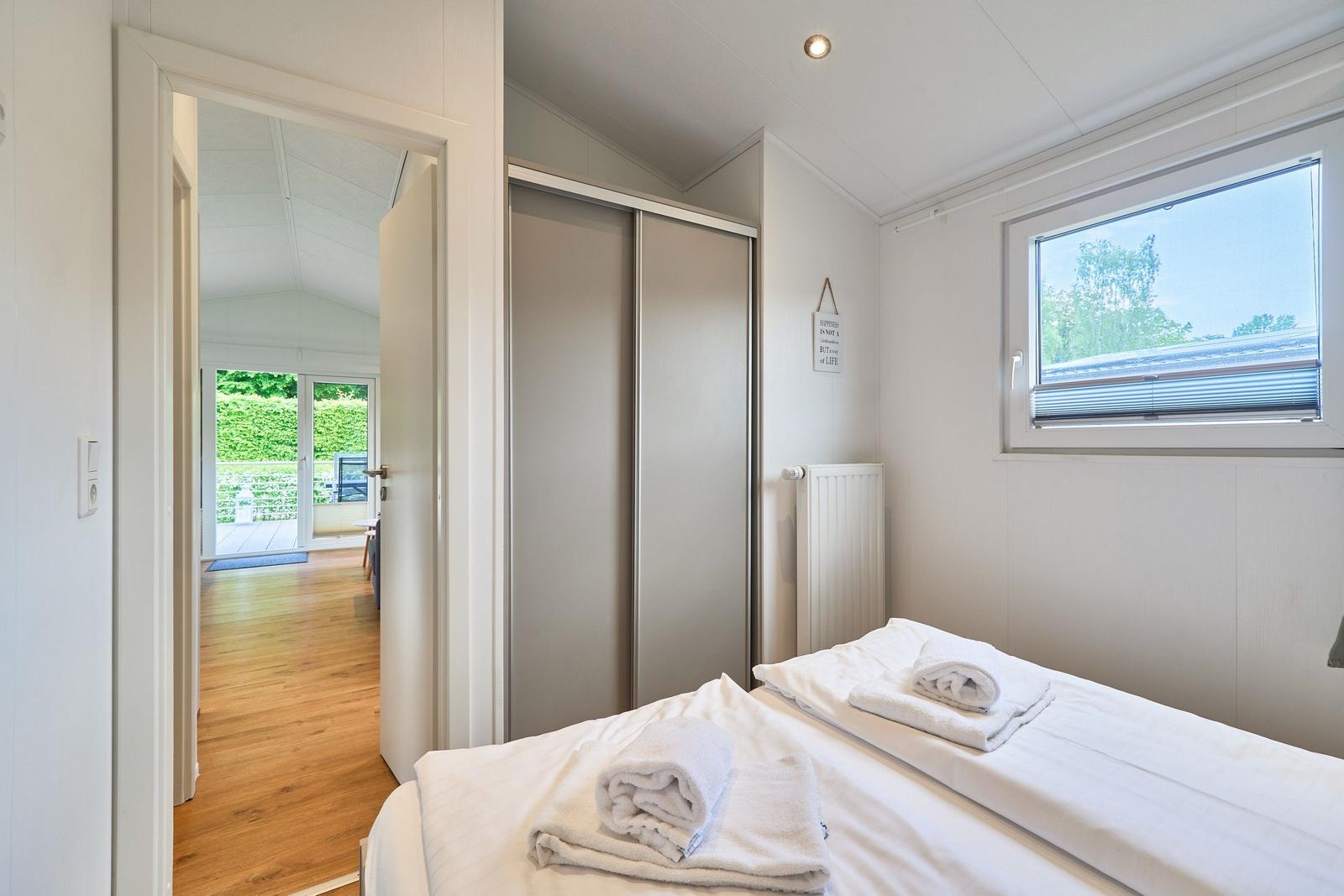Schlafzimmer mit Bett, Kleiderschrank und Fenster. Durchgang zu Wohnbereich mit Holzboden.