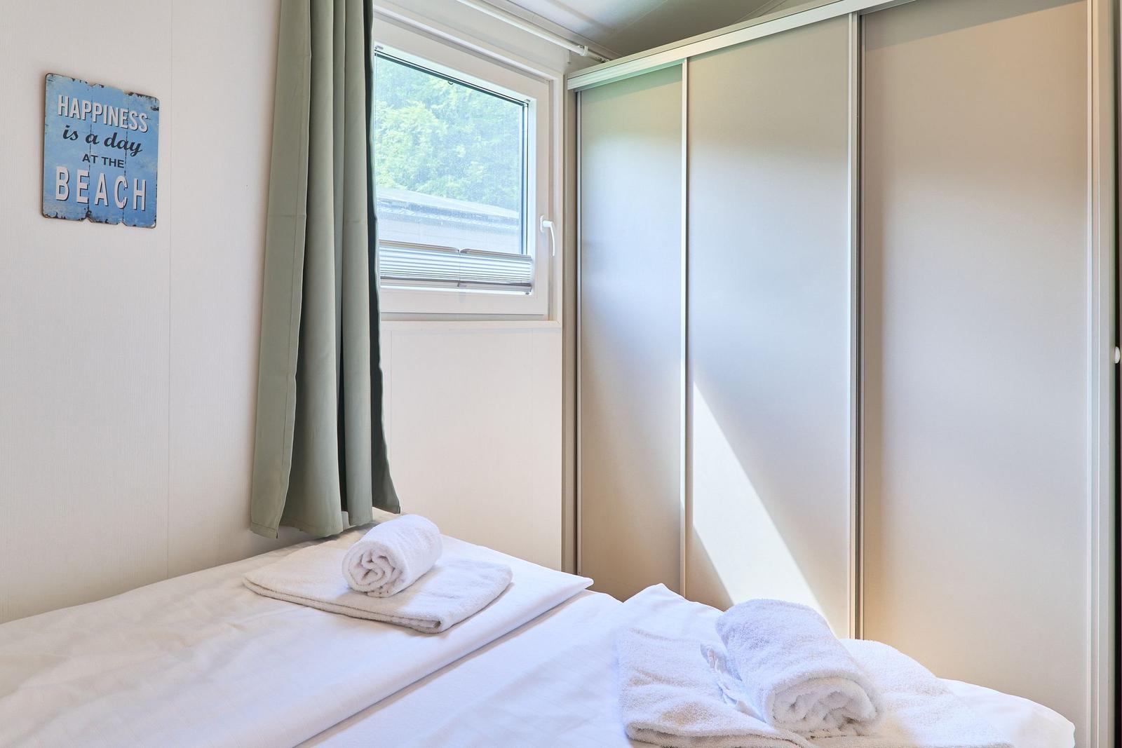 Schlafzimmer mit Bett, Fenster und Kleiderschrank. Auf dem Bett liegen Handtücher.