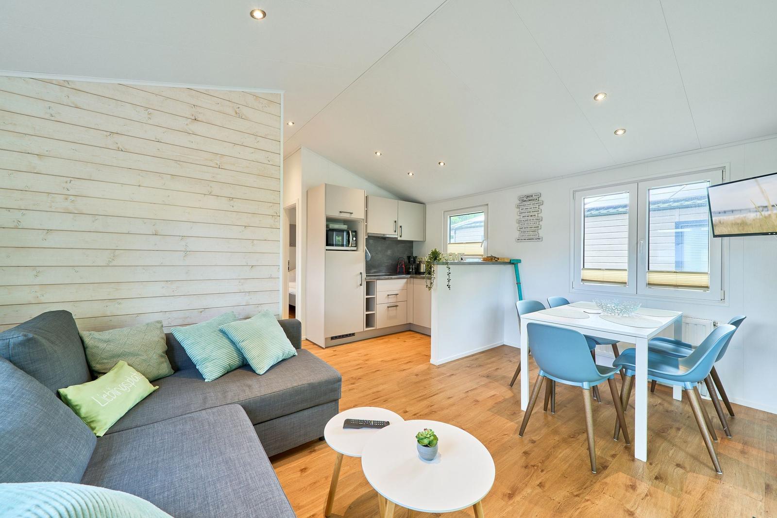 Wohnzimmer mit Sofa, Essbereich und Küchenzeile. Holzwand, Holzboden und Fenster.