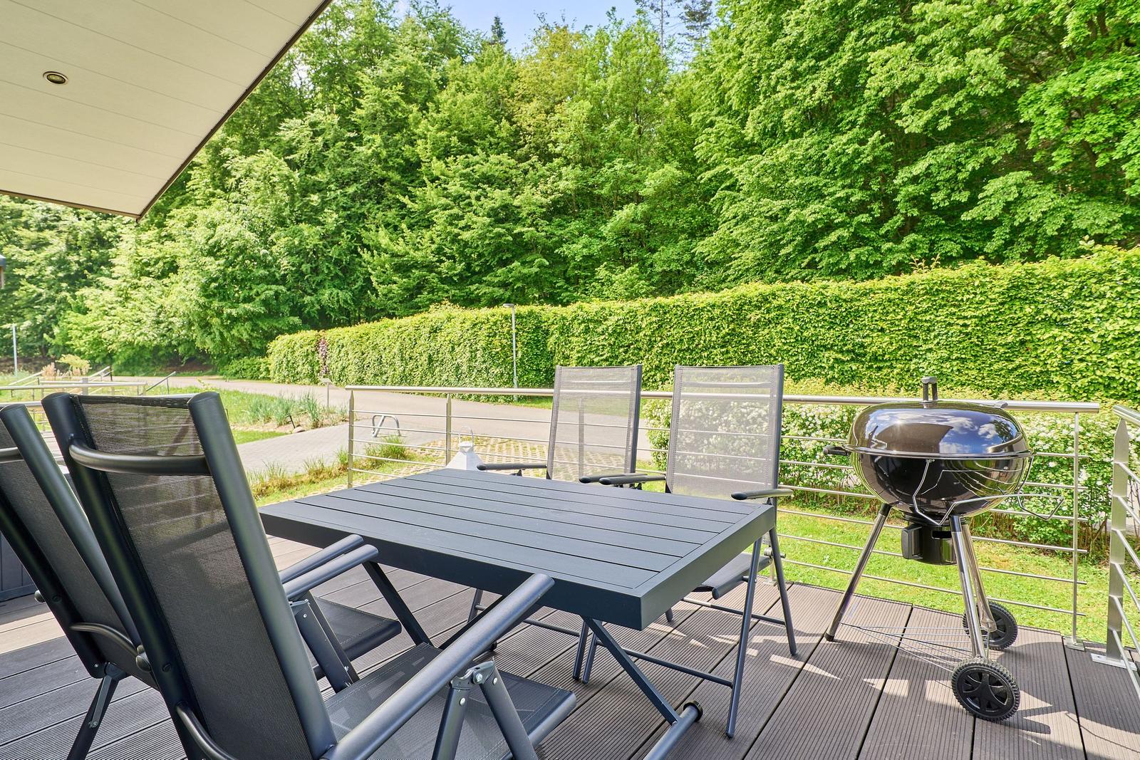 Terrasse mit Tisch, Stühlen und Grill. Hintergrund: grüne Hecke und Wald.