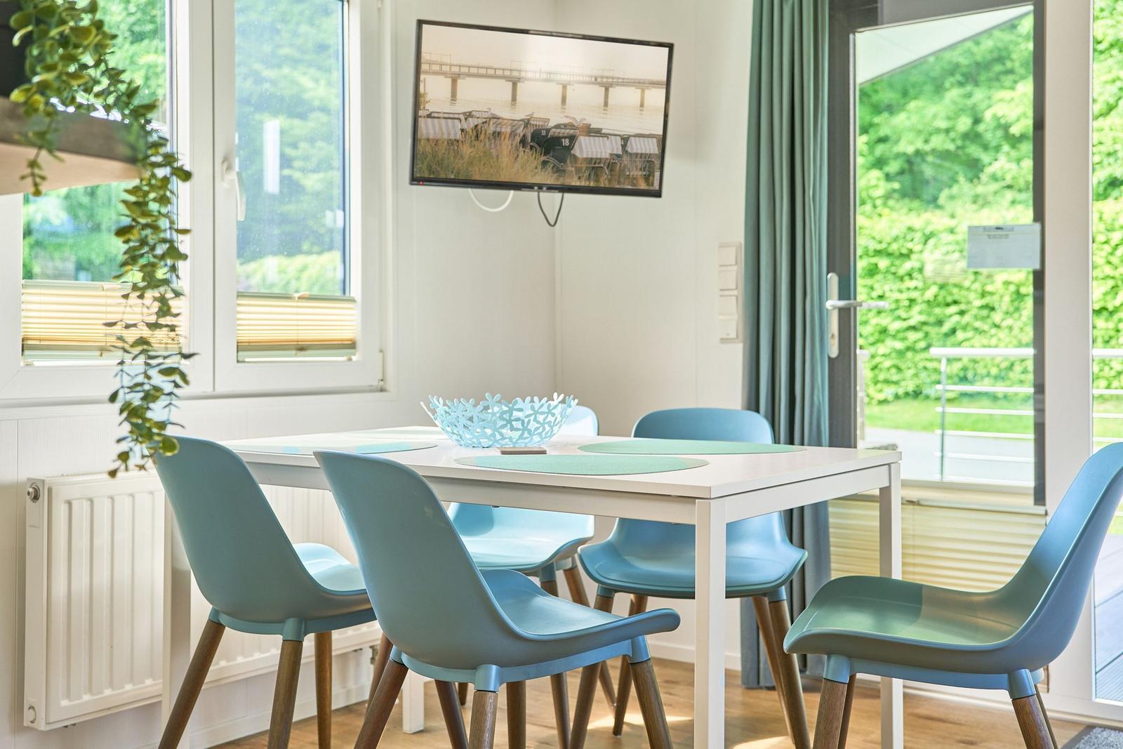 Esstisch mit sechs blauen Stühlen, Fernseher an der Wand, Fenster mit Blick auf Grün.