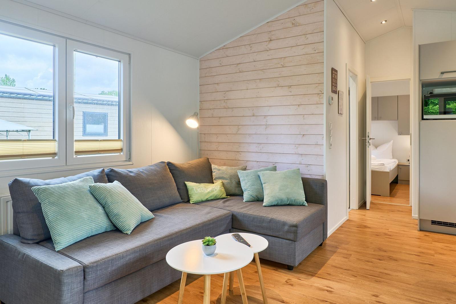 Wohnzimmer mit Sofa, Tisch und Fenster. Im Hintergrund ist ein Schlafzimmer sichtbar.