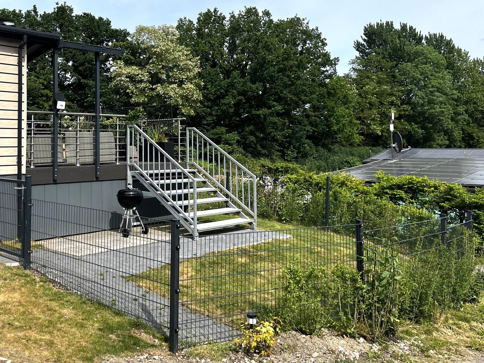 Terrasse mit Stufen, Grill und Gartenzaun. Hintergrund: Bäume und Solaranlage.