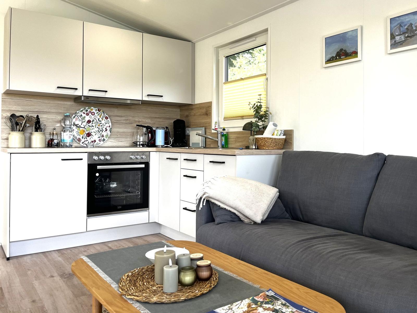 Küche mit Schränken, Herd und Fenster. Wohnbereich mit Sofa und Tisch.