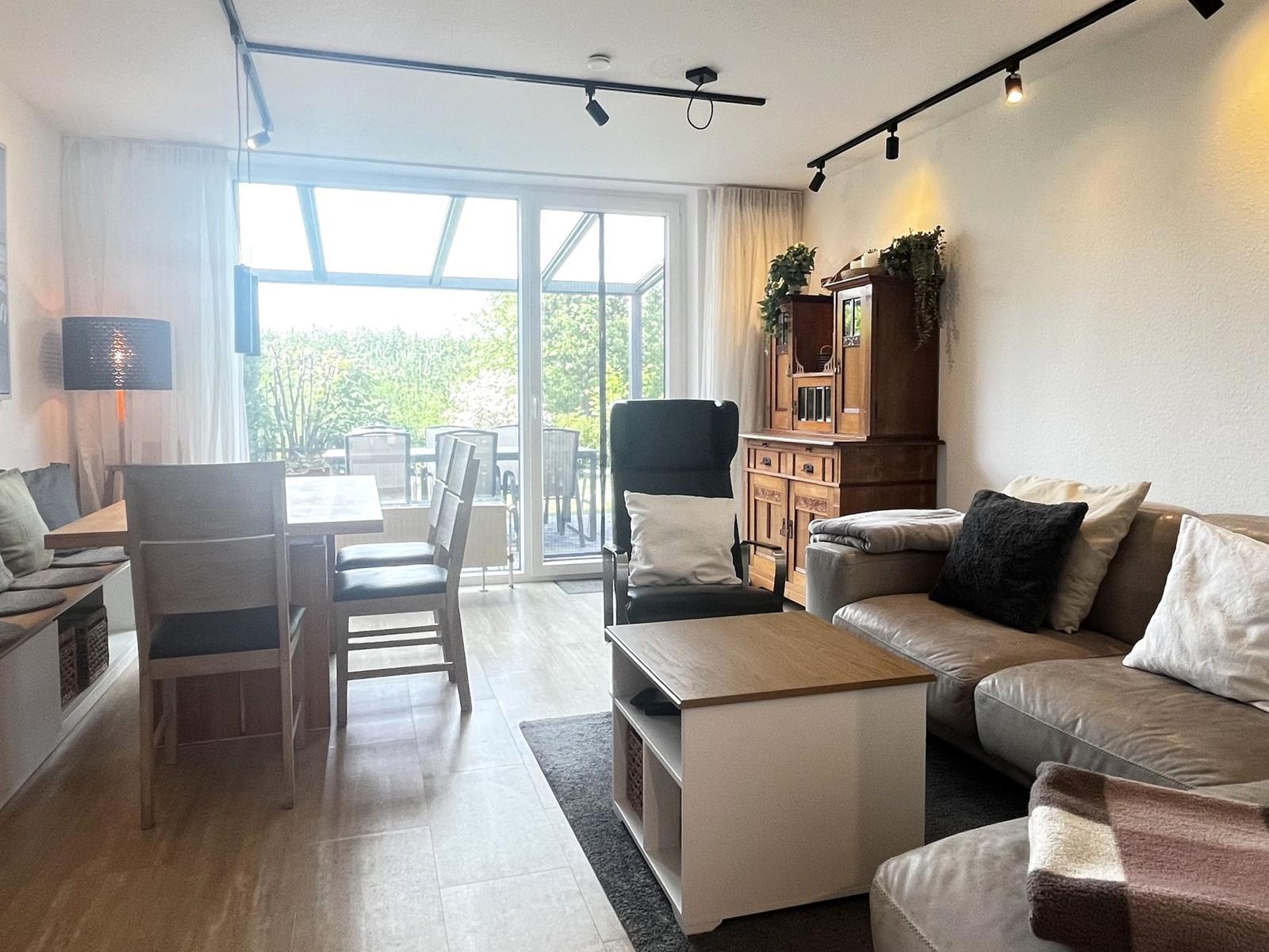 Wohn- und Essbereich mit Sofa, Tisch und Sitzgelegenheiten. Durchgang zum Balkon mit Gartenblick.