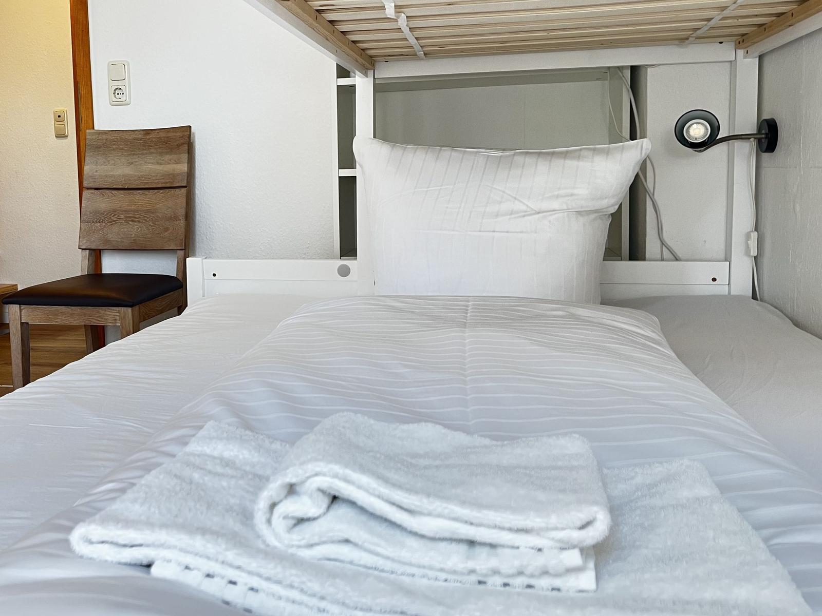 Ein Bett mit weißer Bettwäsche und einer gefalteten Handtuchrolle. Eine Holzstuhllehne ist links sichtbar.