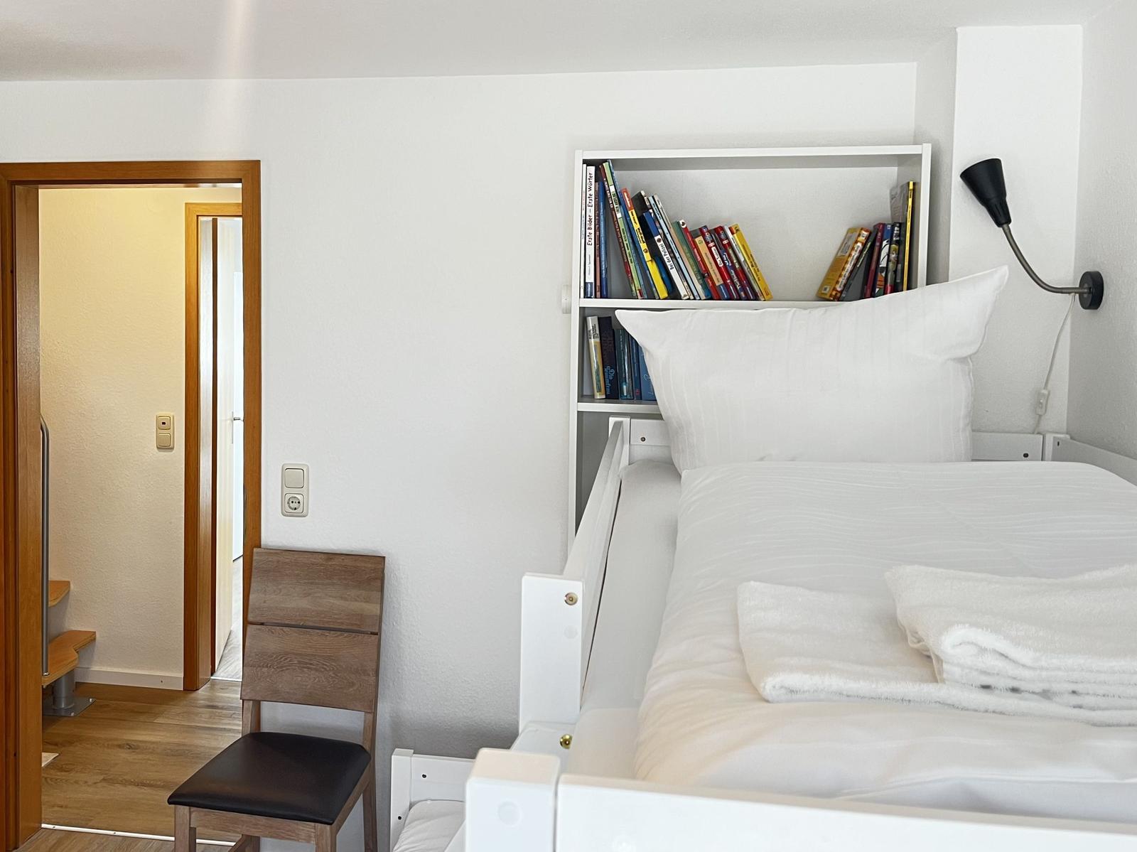 Schlafzimmer mit weißem Bett, Bücherregal und Stuhl. Tür führt in angrenzenden Raum.
