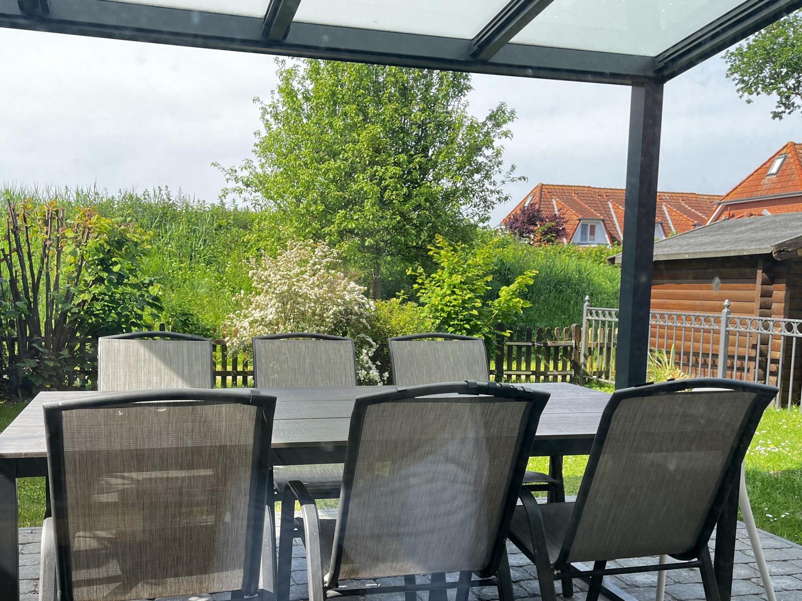 Terrasse mit Tisch und Stühlen unter Glasdach. Grüner Garten und Nachbarhäuser im Hintergrund.