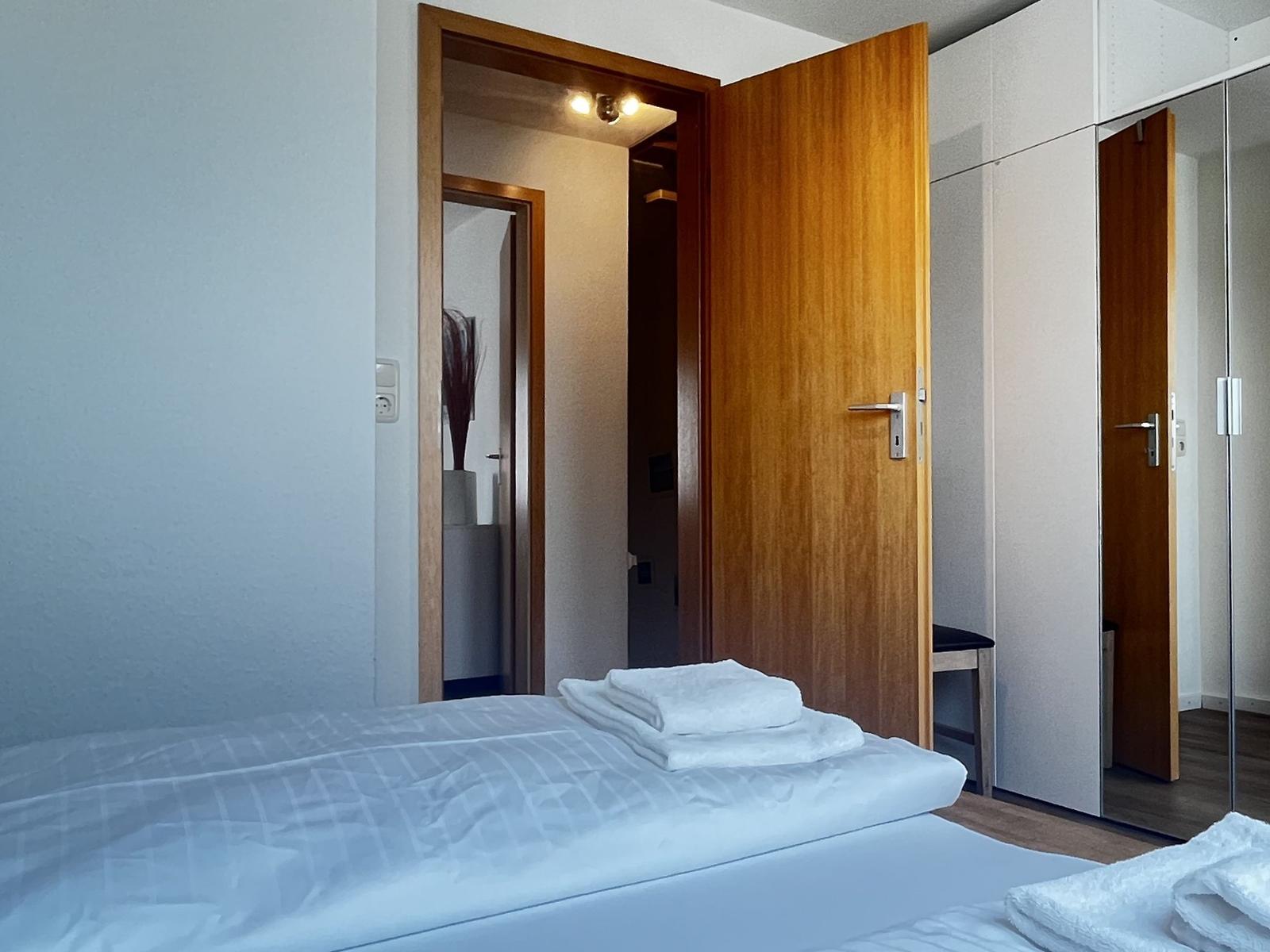 Schlafzimmer mit Bett, Bad und Kleiderschrank. Weiße Wände und Holztüren.