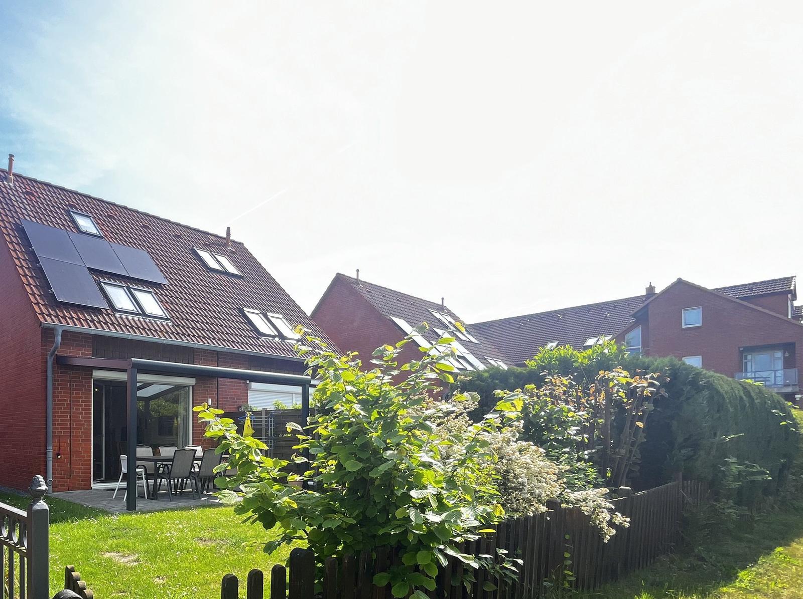 Rotes Haus mit Garten und Terrasse. Solarpaneele auf Dach. Grüne Pflanzen im Vordergrund.