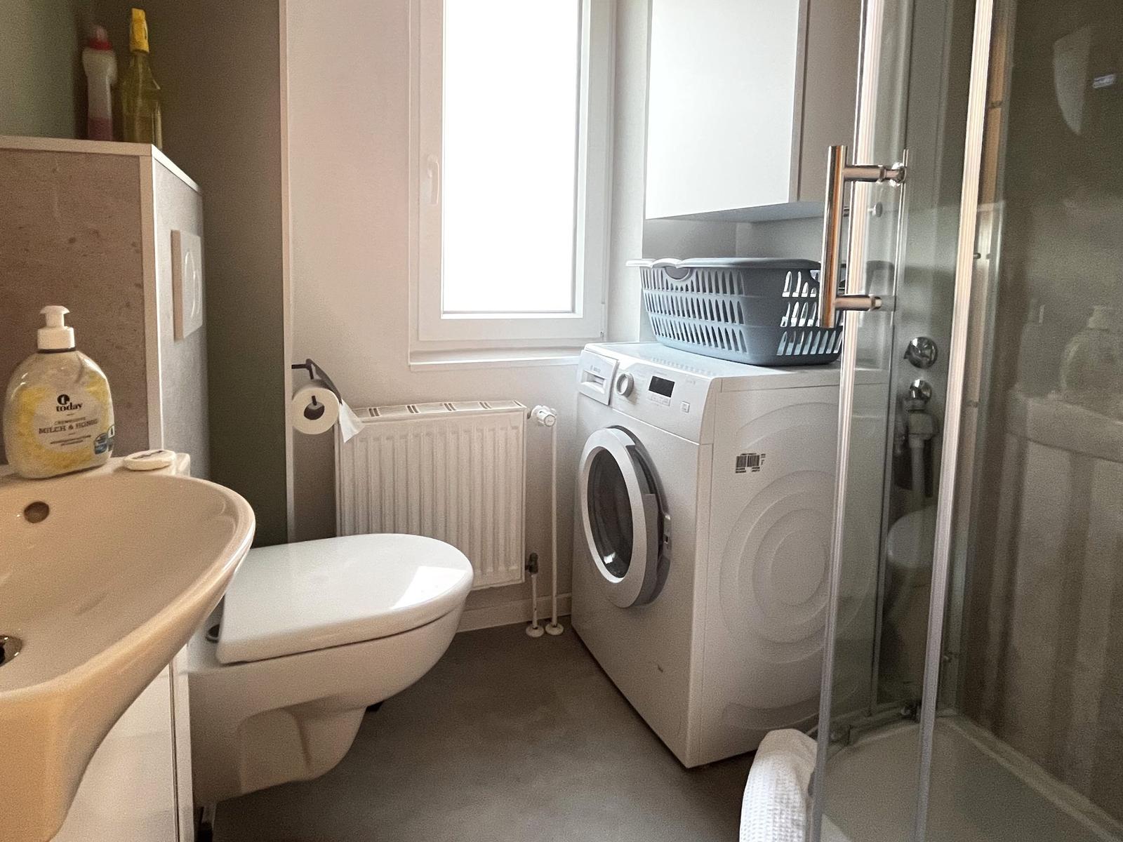 Badezimmer mit Waschmaschine, Dusche und Toilette. Fenster sorgt für Tageslicht.