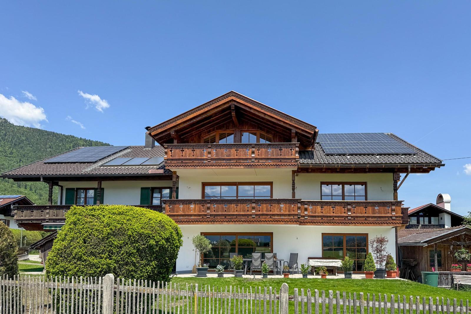 Haus mit Balkonen, Solaranlagen und Garten. Berglandschaft im Hintergrund.