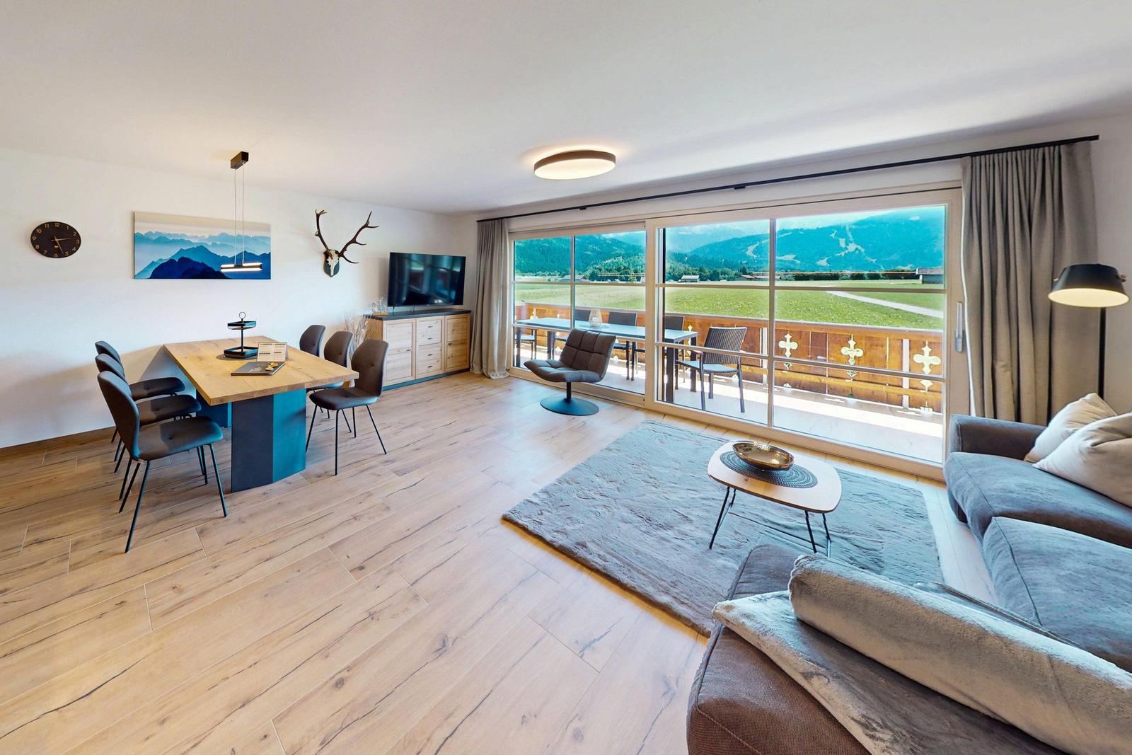 Wohn-Essbereich mit Balkon und Bergblick. Holzboden, Sofa, Esstisch und TV.