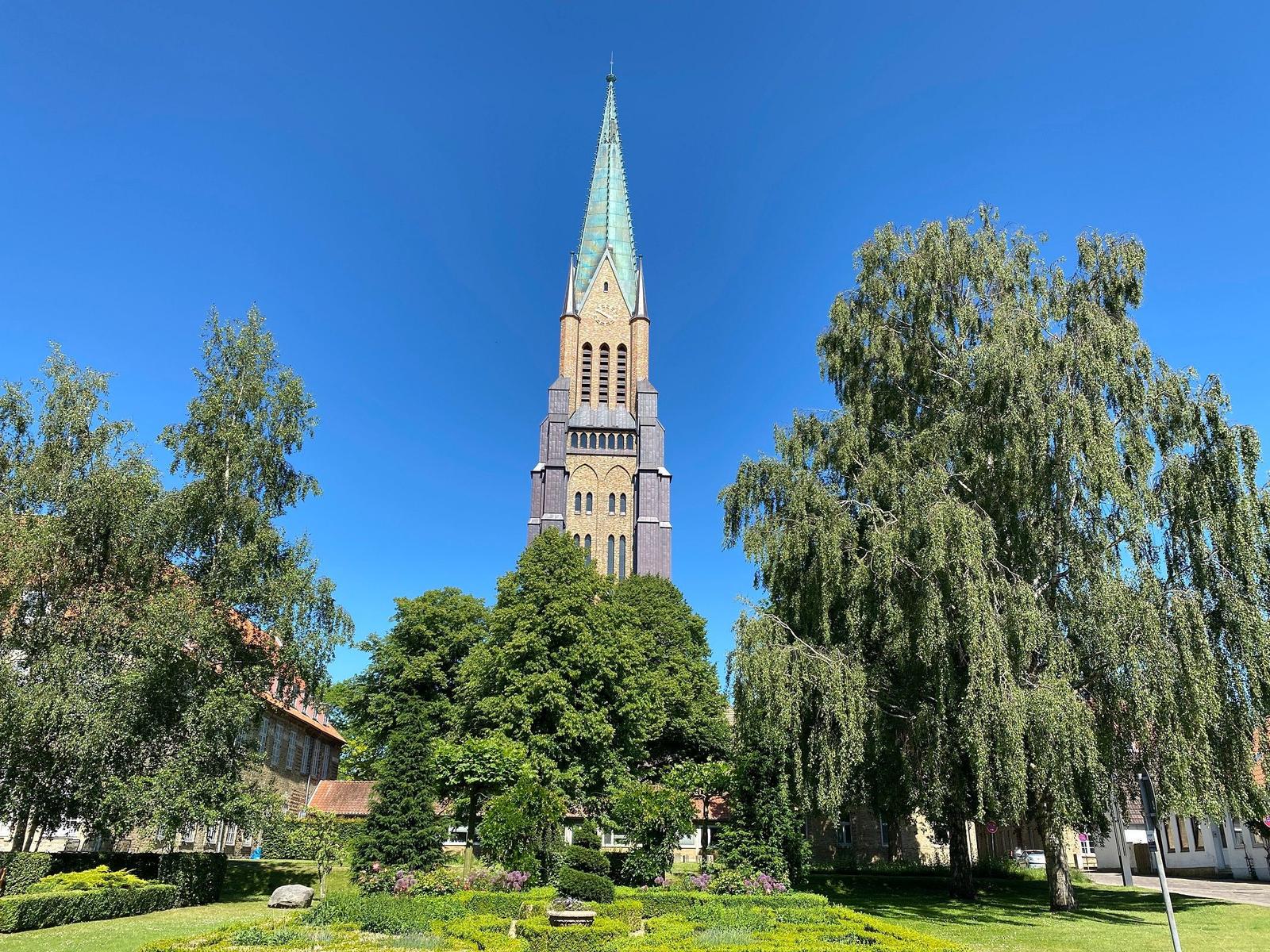 Hoch aufragender Kirchturm mit grünem Spitzdach, umgeben von Bäumen und Rasen.