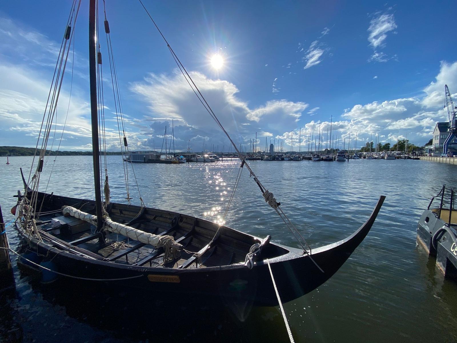 Ein schwarzes Segelboot liegt im Hafen. Im Hintergrund sind weitere Boote und ein Himmel mit Sonne und Wolken zu sehen.