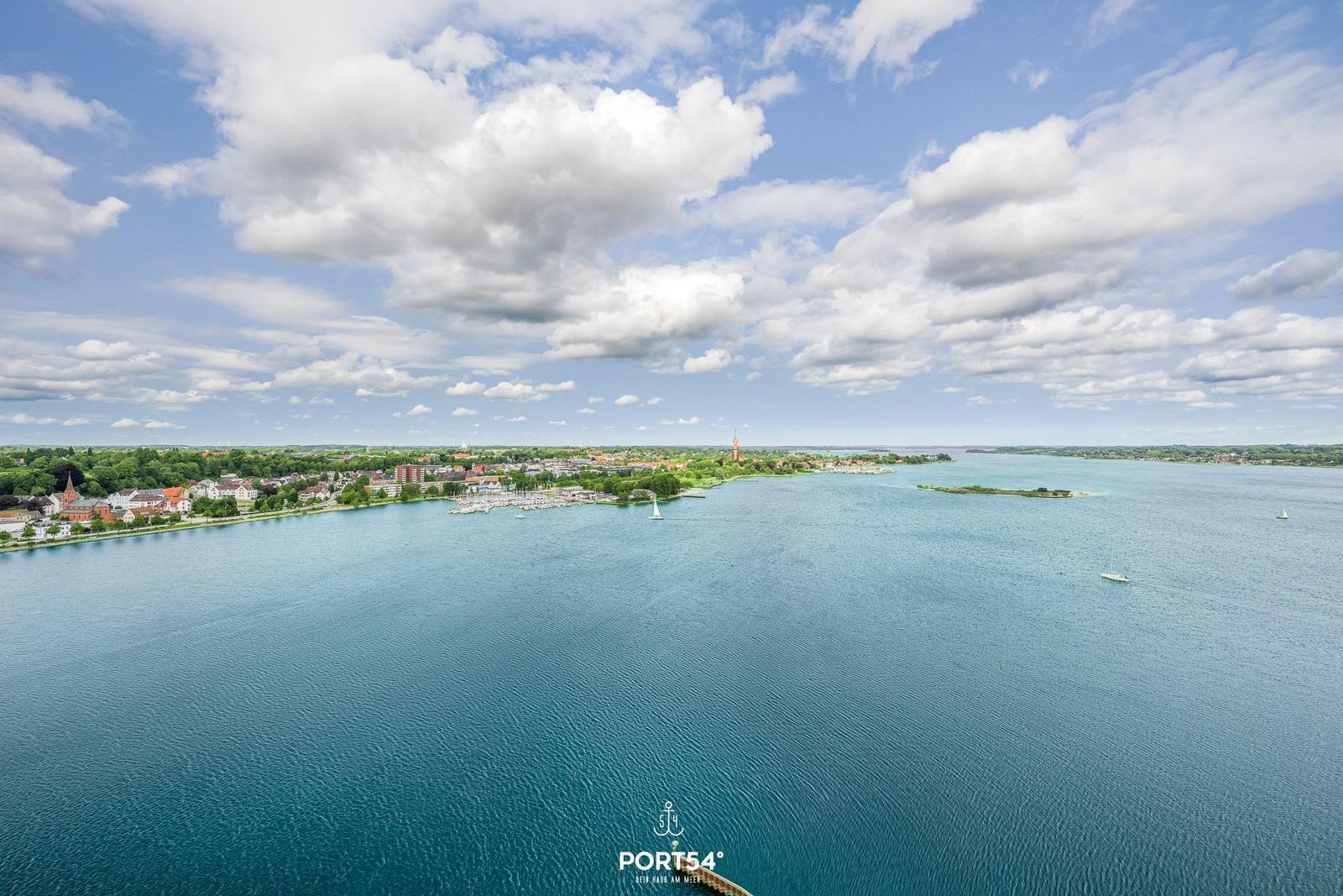 Weitläufiger Blick auf einen Hafen mit Booten und Stadt am Ufer unter blauem Himmel mit Wolken.