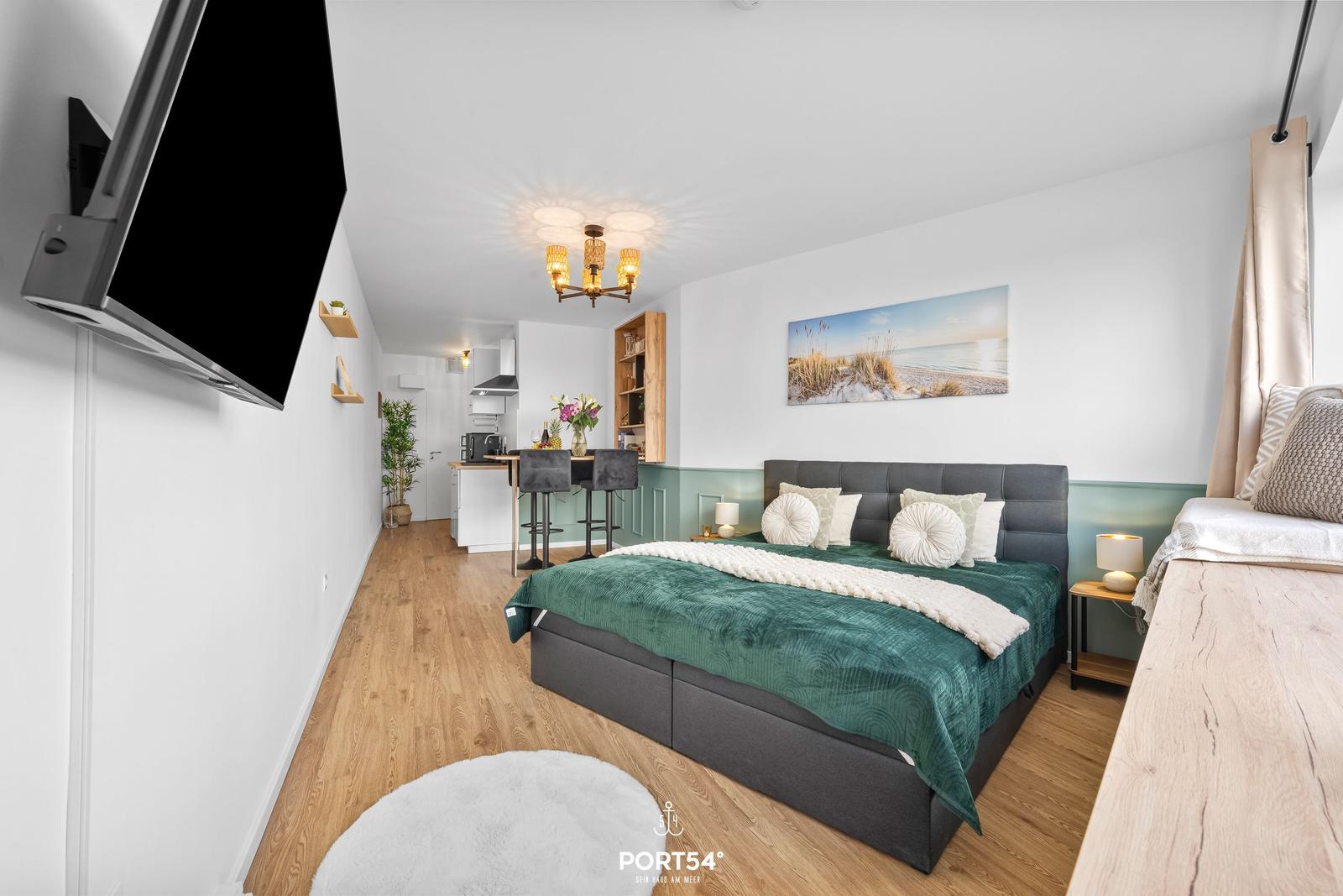 Schlafzimmer mit Bett, TV und Küchenbereich im Hintergrund.