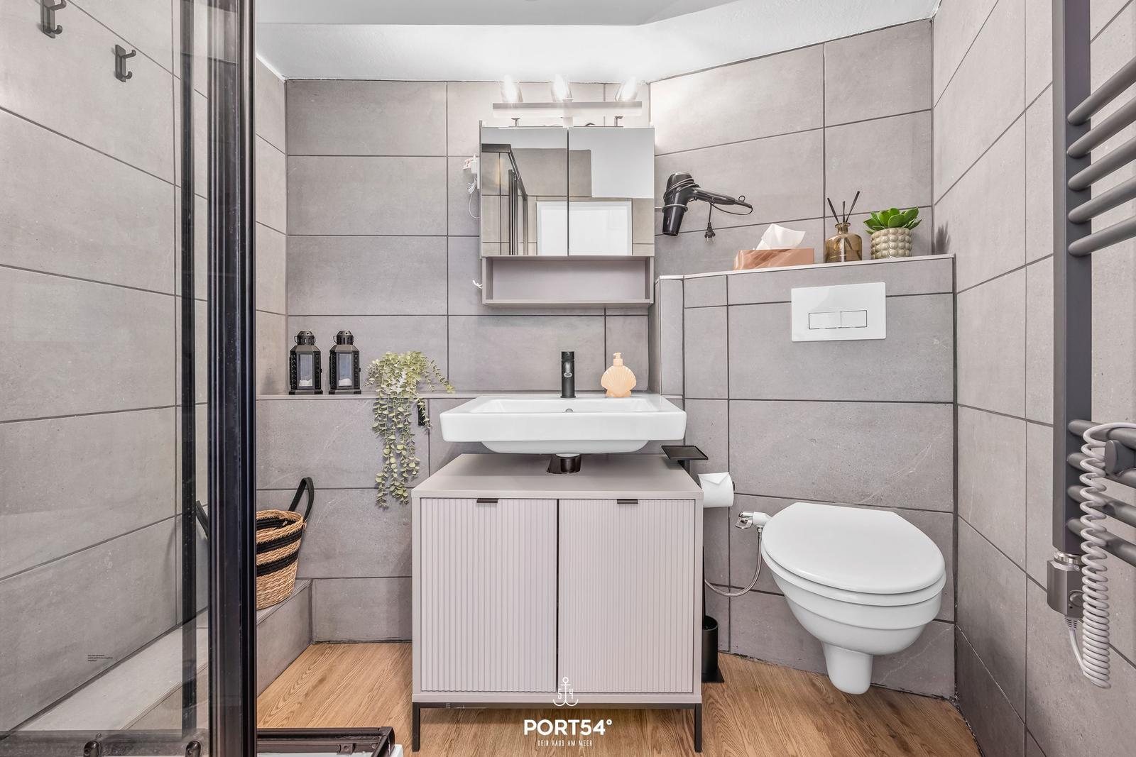 Badezimmer mit Dusche, Waschbecken und Toilette. Graue Fliesen und Holzboden.