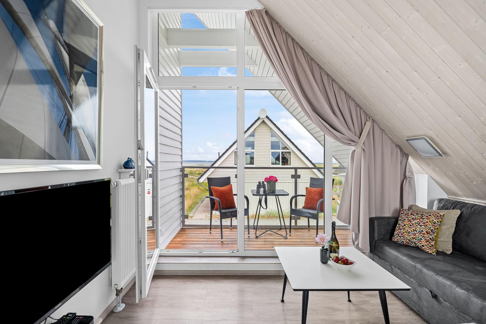 Wohnbereich mit Balkon, Sofa und Fernseher. Blick auf Haus und Strand.