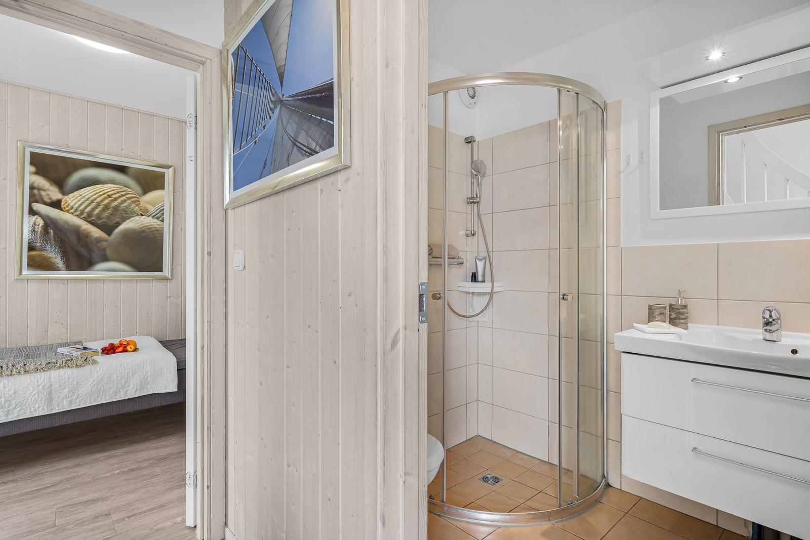 Badezimmer mit Dusche, Waschbecken und Spiegel. Blick in Schlafzimmer mit Bett und Wandbild.