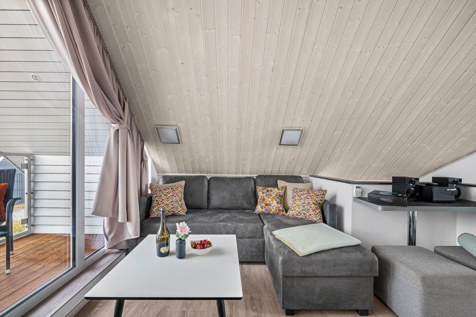 Wohnbereich mit grauem Sofa, Couchtisch und Holzdecke. Große Fenster mit Blick auf Balkon.