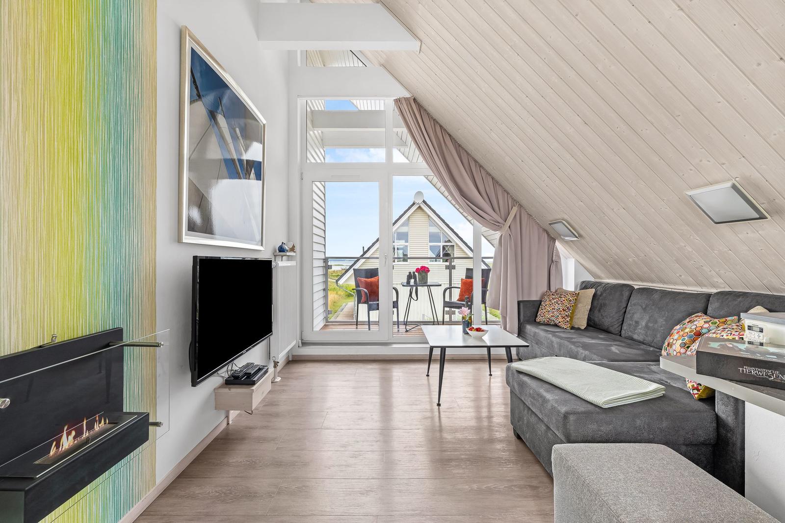 Wohnzimmer mit Sofa, Fernseher und Kamin. Durchgang zum Balkon mit Sicht auf Haus und Meer.