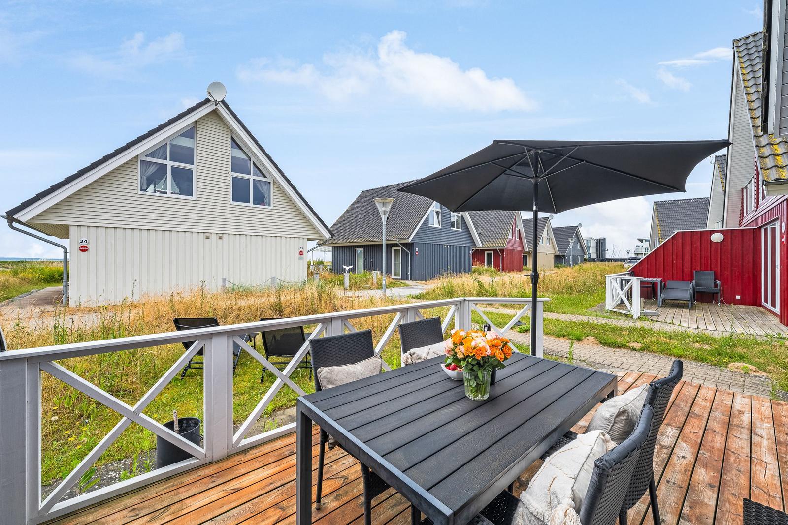 Terrasse mit Tisch, Stühlen und Sonnenschirm. Hintergrund: mehrere Häuser und Grasland.