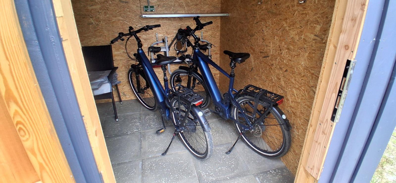 Zwei blaue E-Bikes stehen in einem Holzraum mit Steinboden und Stuhl.