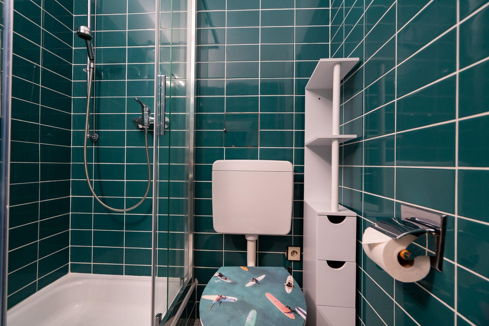 Badezimmer mit Dusche, WC und Regal an grünen Fliesen.
