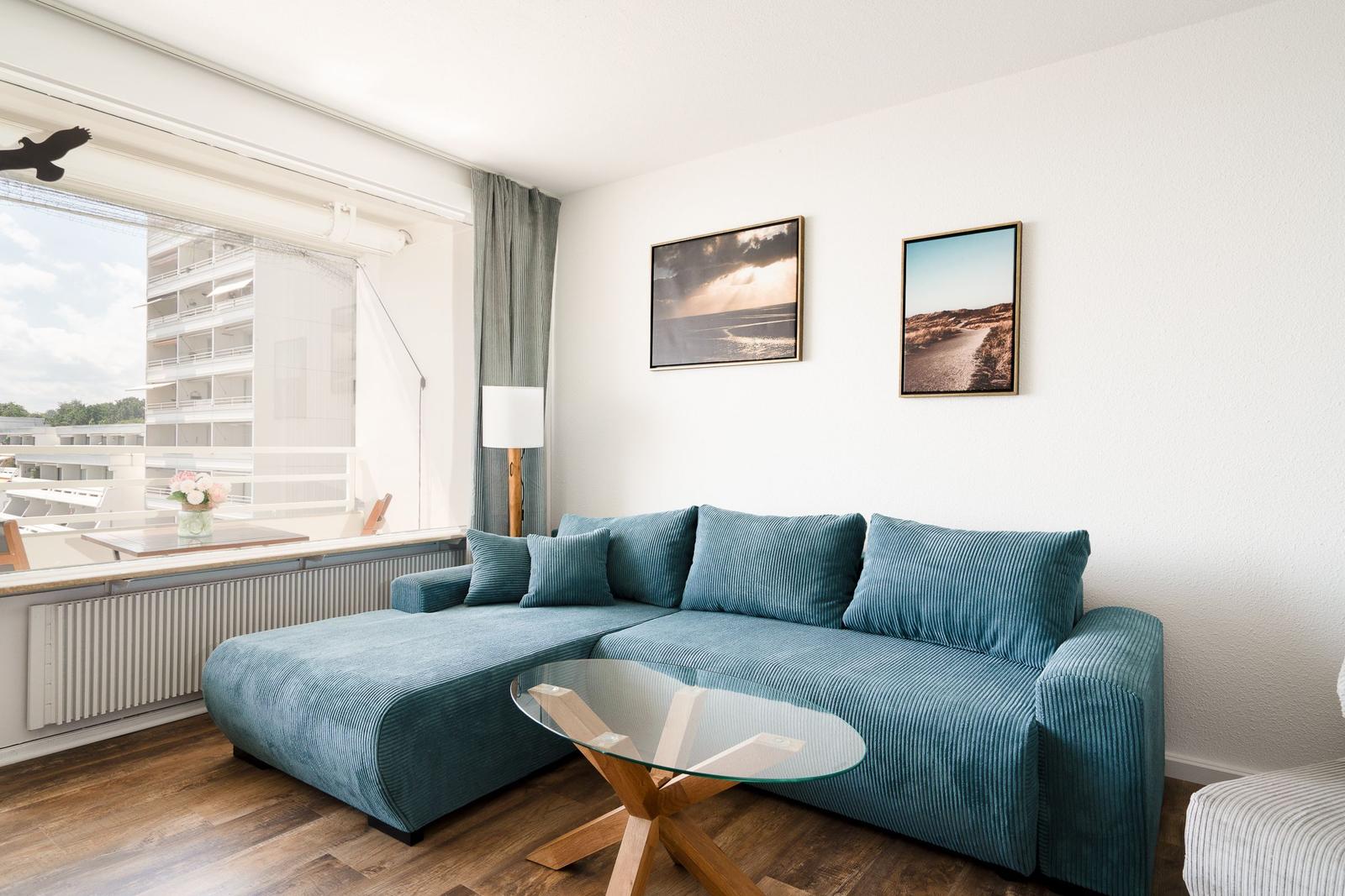 Ein Wohnzimmer mit blauem Sofa, Glas-Tisch und Fensterblick auf Gebäude.