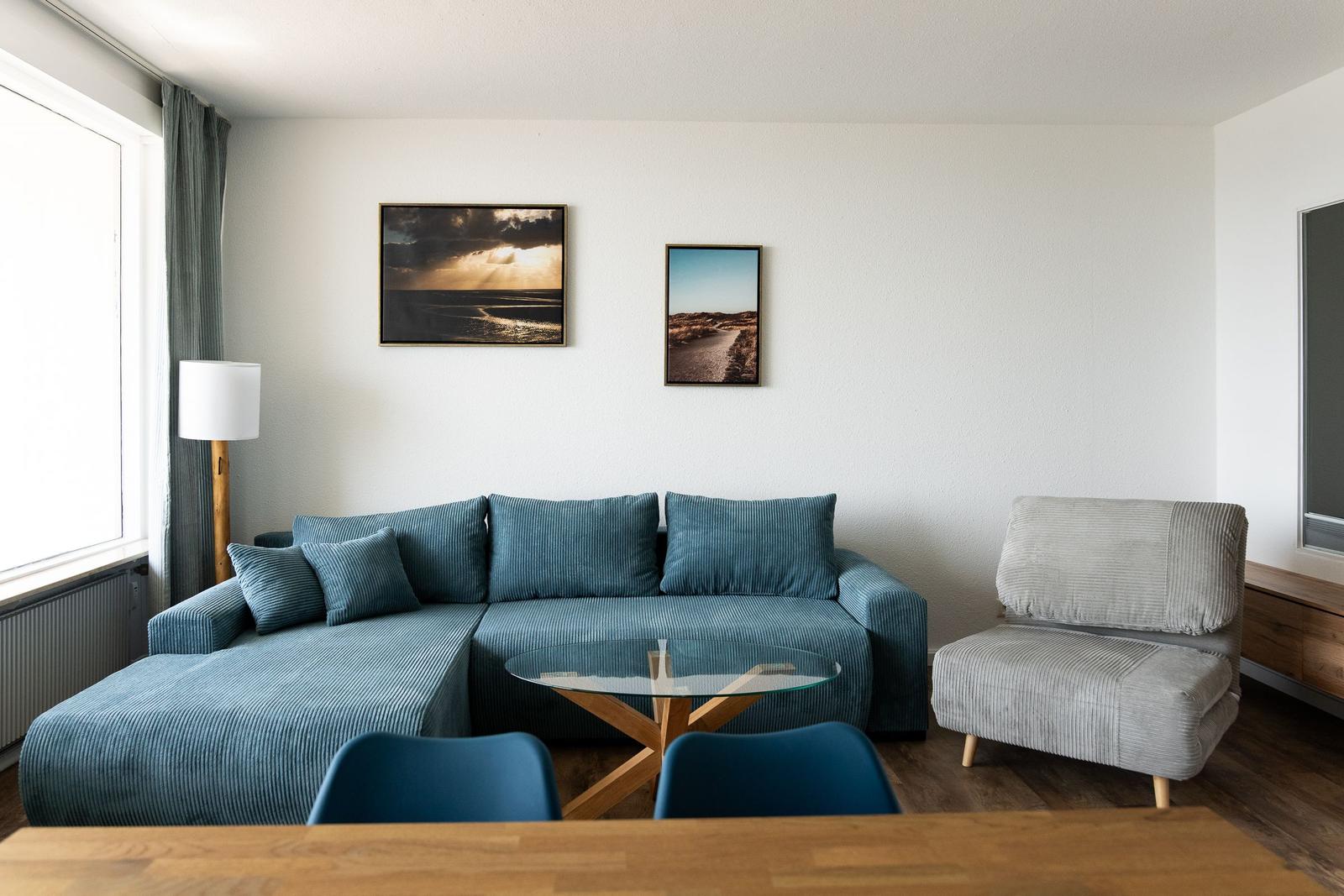 Wohnzimmer mit blauem Sofa, Glas-Tisch und Wandbildern.