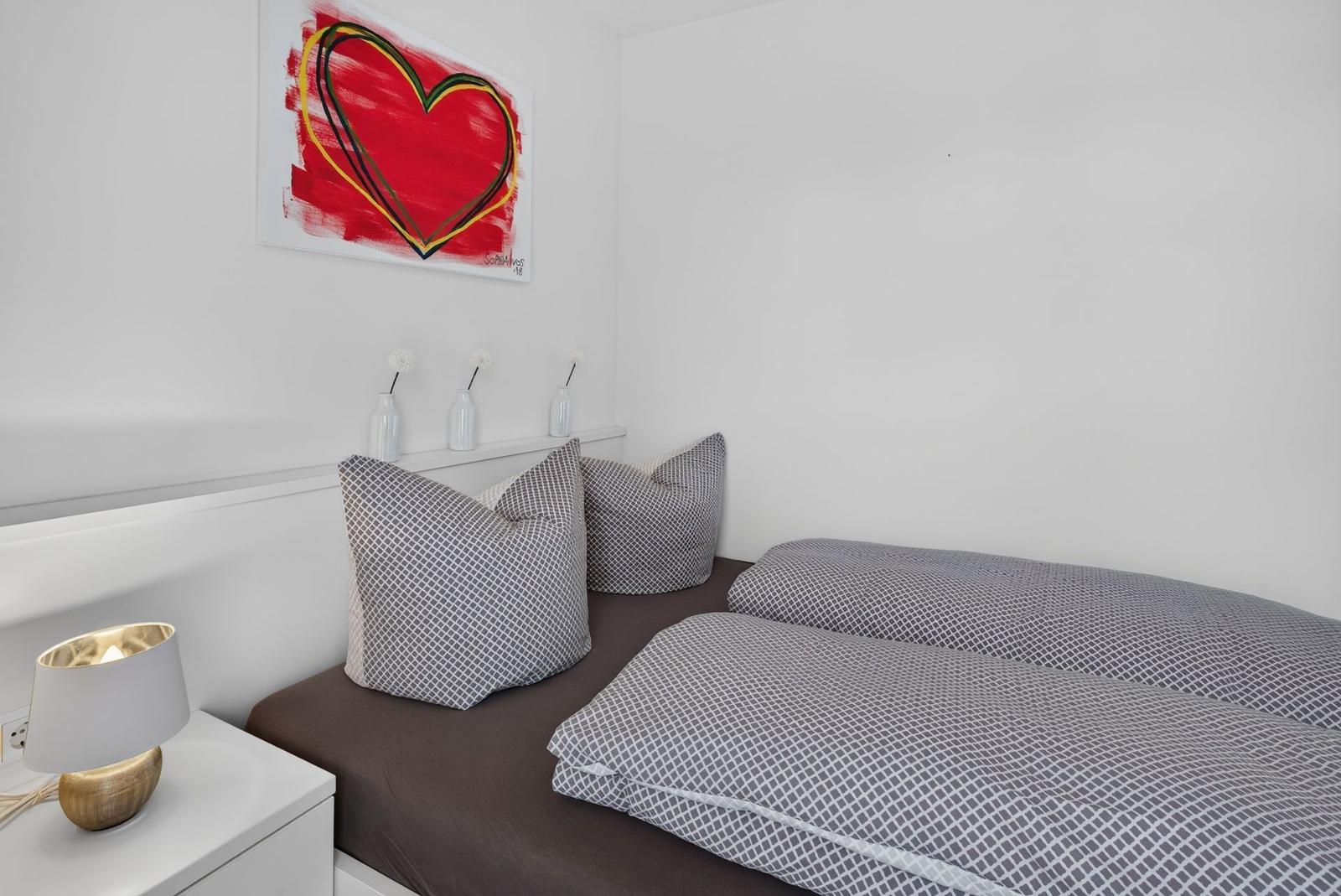 Schlafzimmer mit Bett, Kissen und Wandbild. Nachttisch mit Lampe daneben.
