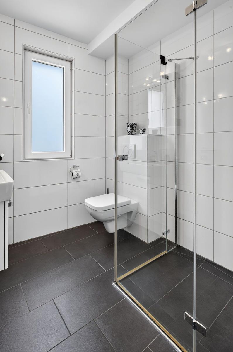 Badezimmer mit Dusche, Toilette und Fenster. Weiße Wände, dunkler Boden.