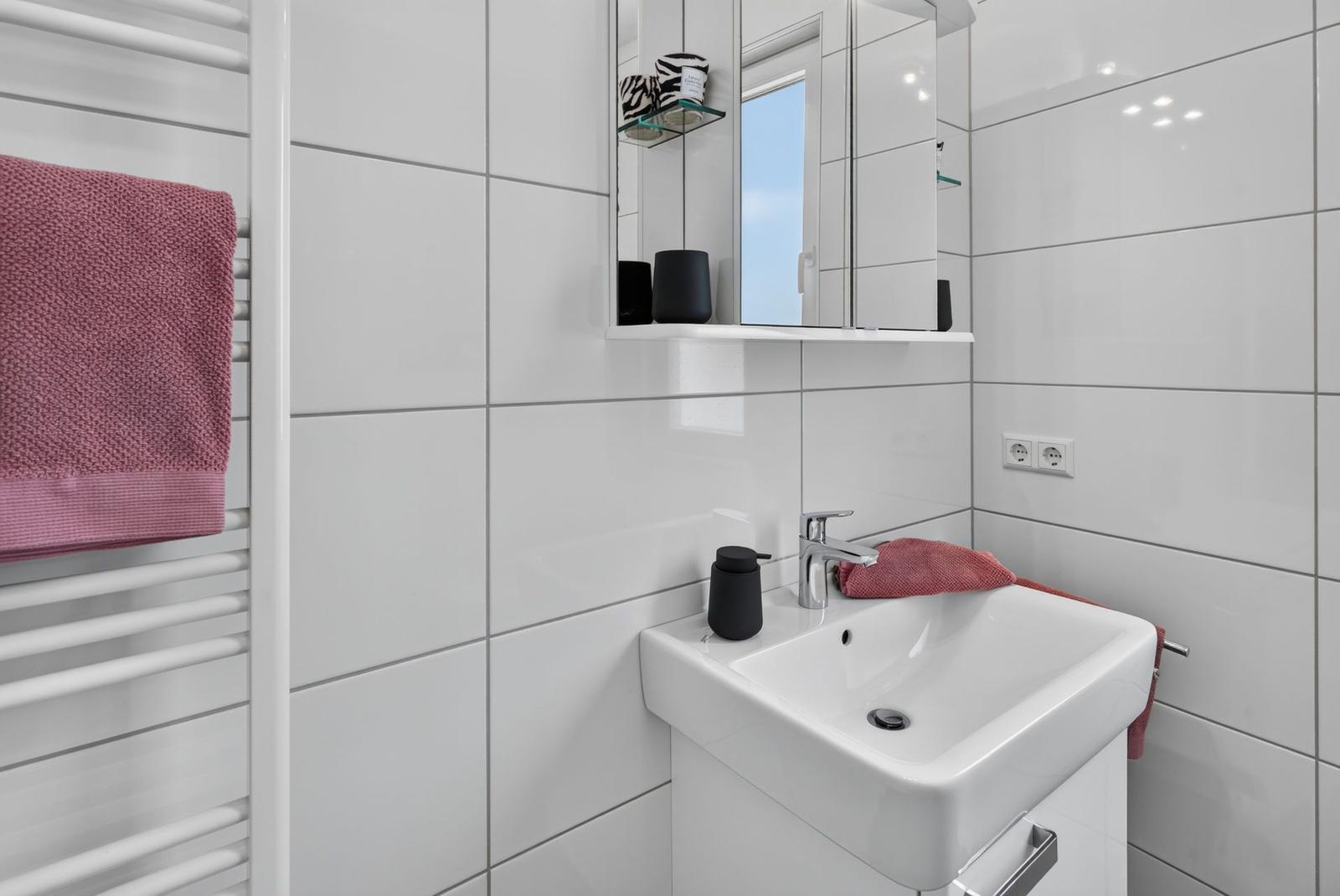 Badezimmer mit weißer Fliesenwand, Waschbecken, Spiegel und Handtuchhalter.