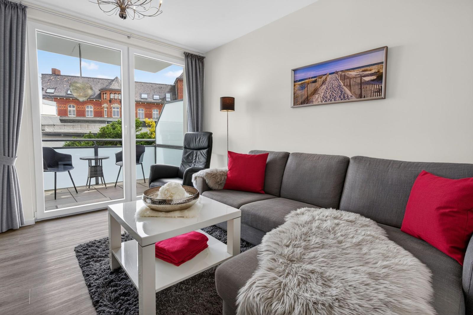 Wohnzimmer mit grauem Sofa, roten Kissen und Balkon mit Blick auf Gebäude.