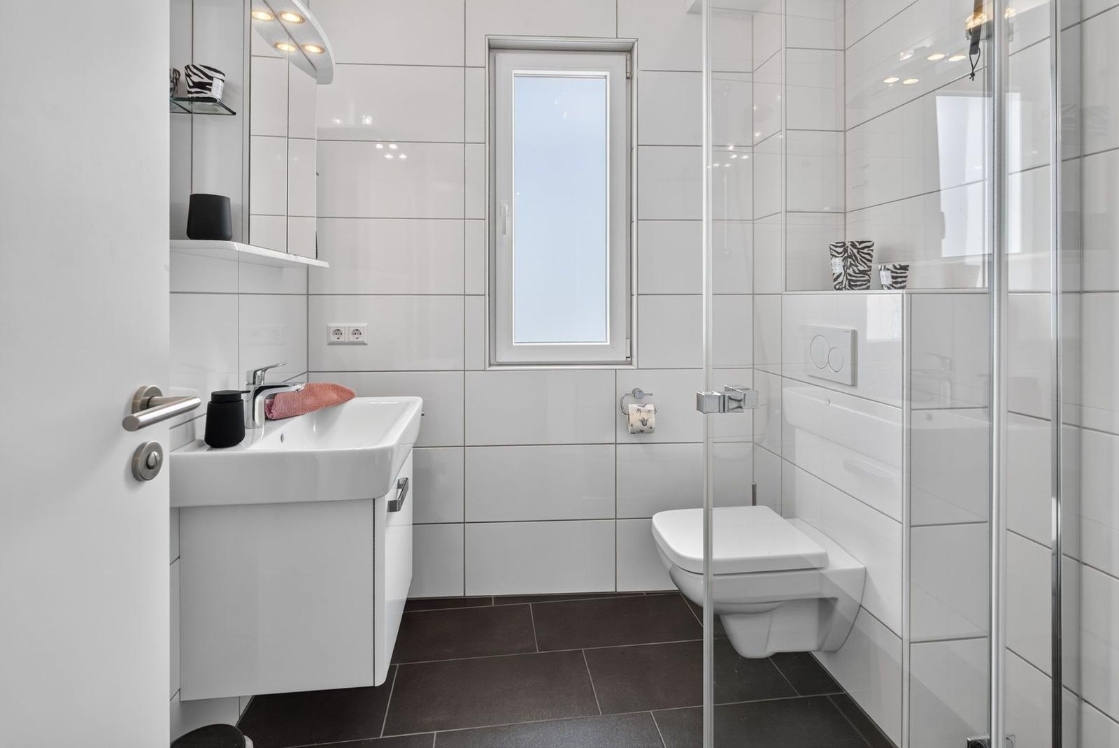 Badezimmer mit Dusche, Toilette und Waschbecken. Weiße Fliesen und dunkle Bodenfliesen.