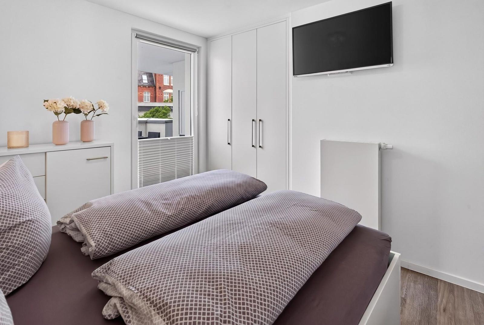 Schlafzimmer mit Bett, Kleiderschrank und TV. Fenster mit Blick auf Gebäude.