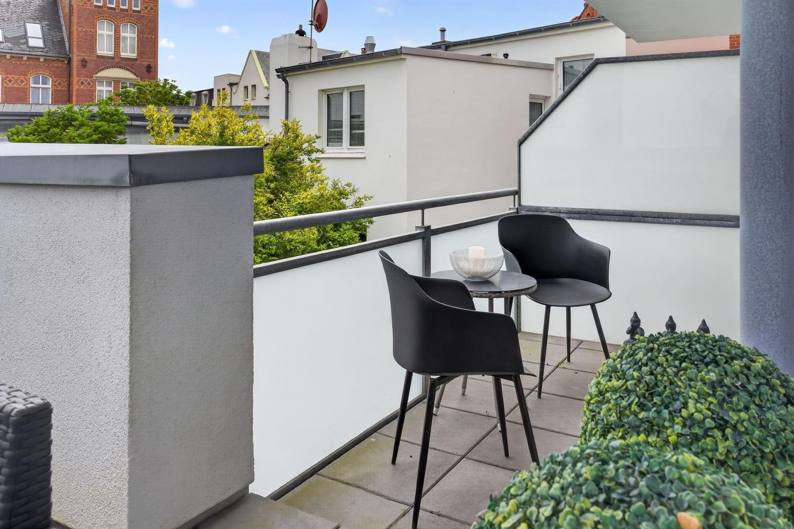Terrasse mit zwei schwarzen Stühlen und Tisch. Blick auf Gebäude und Bäume.